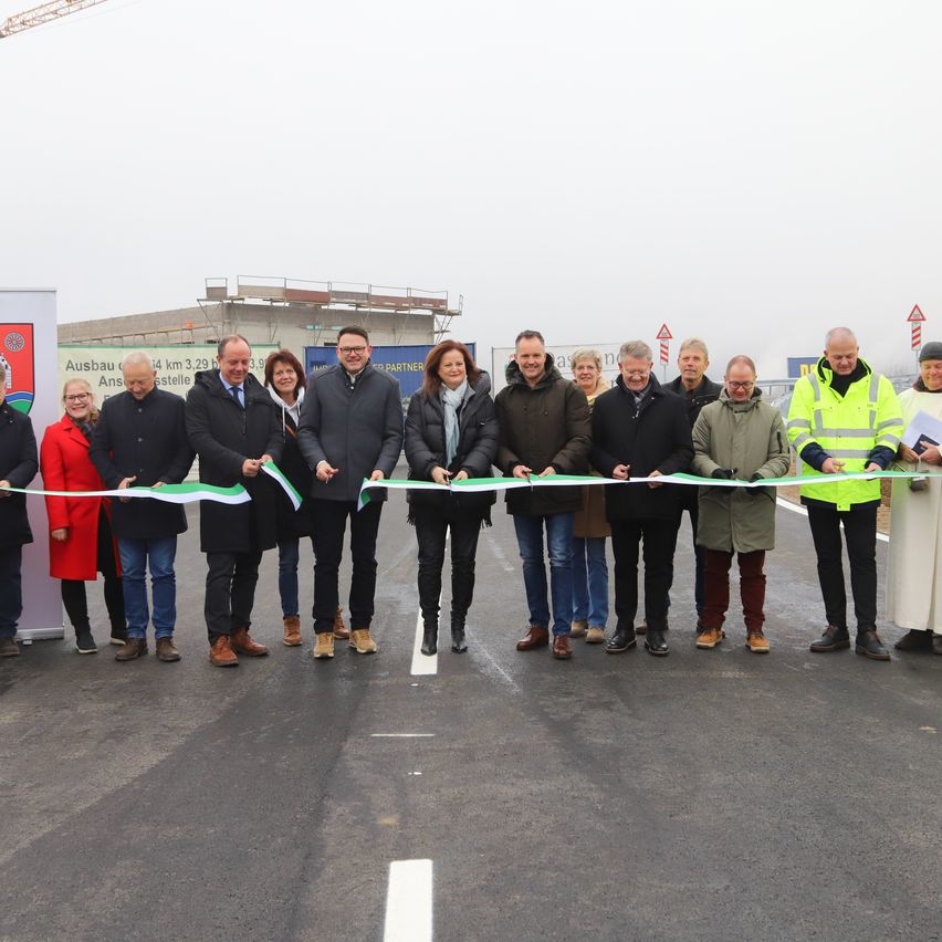 Eine Gruppe von Menschen in Winterkleidung steht vor einer Baustelle und schneidet ein Band mit grünen, weißen und roten Bändern. Hinter ihnen ist ein Gebäude im Bau zu sehen.