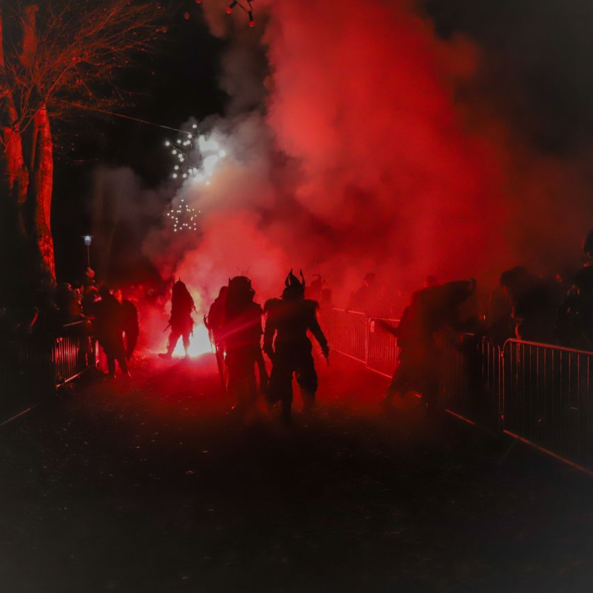 Eine Gruppe von Menschen in Kostümen versammelt sich nachts um ein Lagerfeuer, mit rotem Rauch und Feuerwerk in der Luft.