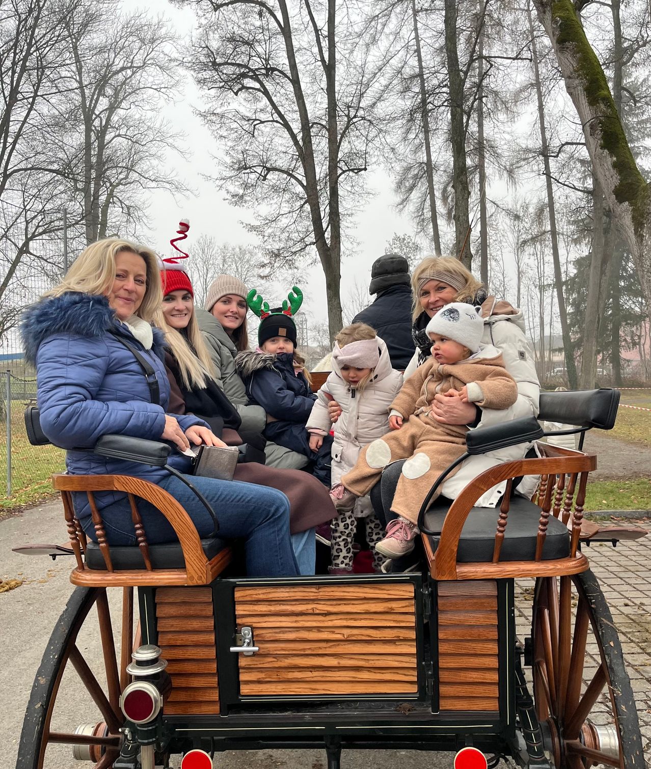 Eine Gruppe von Menschen fährt in einer alten Kutsche, wobei Erwachsene und Kinder Winterkleidung tragen und einige mit Weihnachtsdekorationen. Sie lächeln und scheinen die Fahrt zu genießen. Die Umgebung ist im Freien mit Bäumen im Hintergrund.