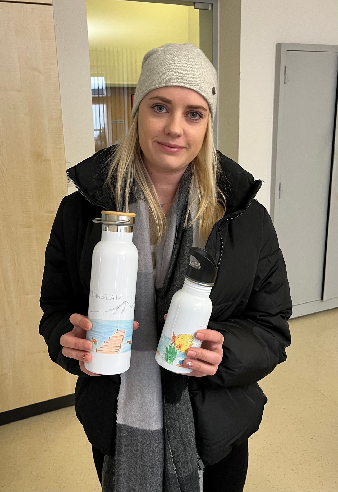 Eine Frau in einem Wintermantel hält zwei weiße Flaschen mit Designs. Die linke Flasche hat ein Berg- und Brückendesign, die rechte ein Fischdesign. Sie ist drinnen in der Nähe einer Holzwand.