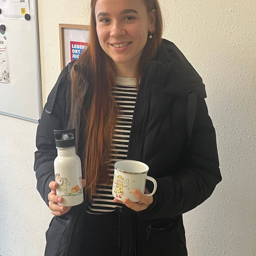 Eine Frau in einer schwarzen Jacke und gestreiftem Shirt hält einen weißen Becher und eine weiße Flasche mit Cartoon-Designs. Sie lächelt und steht vor einer weißen Wand.