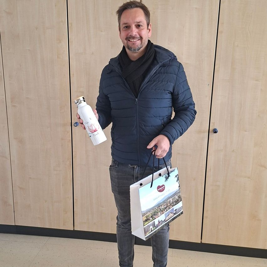 Ein Mann mit einer blauen Jacke und Jeans hält eine weiße Flasche und einen weißen Einkaufsbeutel mit Bildern einer Landschaft und Gebäude vor Holzschränken.