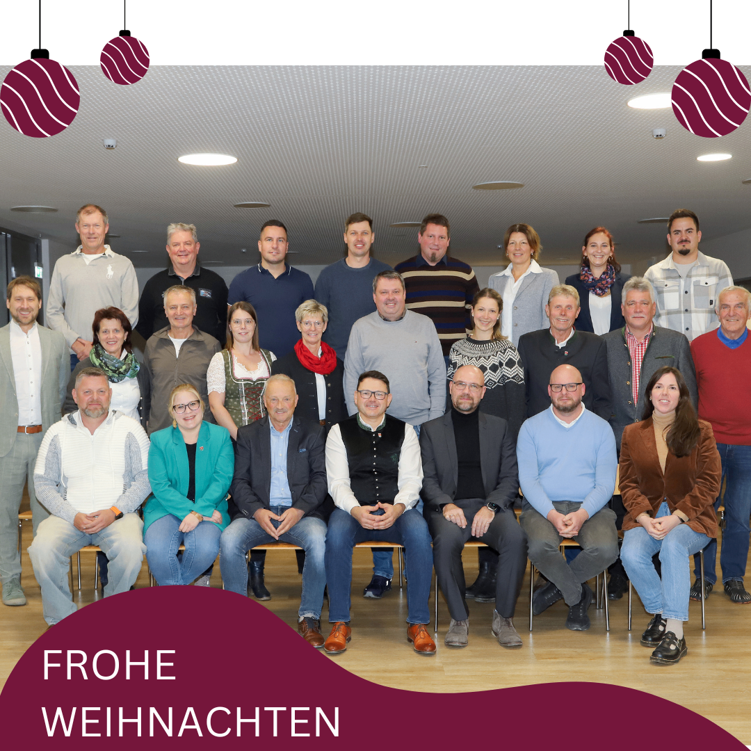 Eine Gruppe von Menschen posiert für ein Foto. Sie sitzen alle auf einer Bank in einem Raum. Sie tragen formelle Kleidung. Der Text 'FROHE WEIHNACHTEN' steht in der unteren linken Ecke.