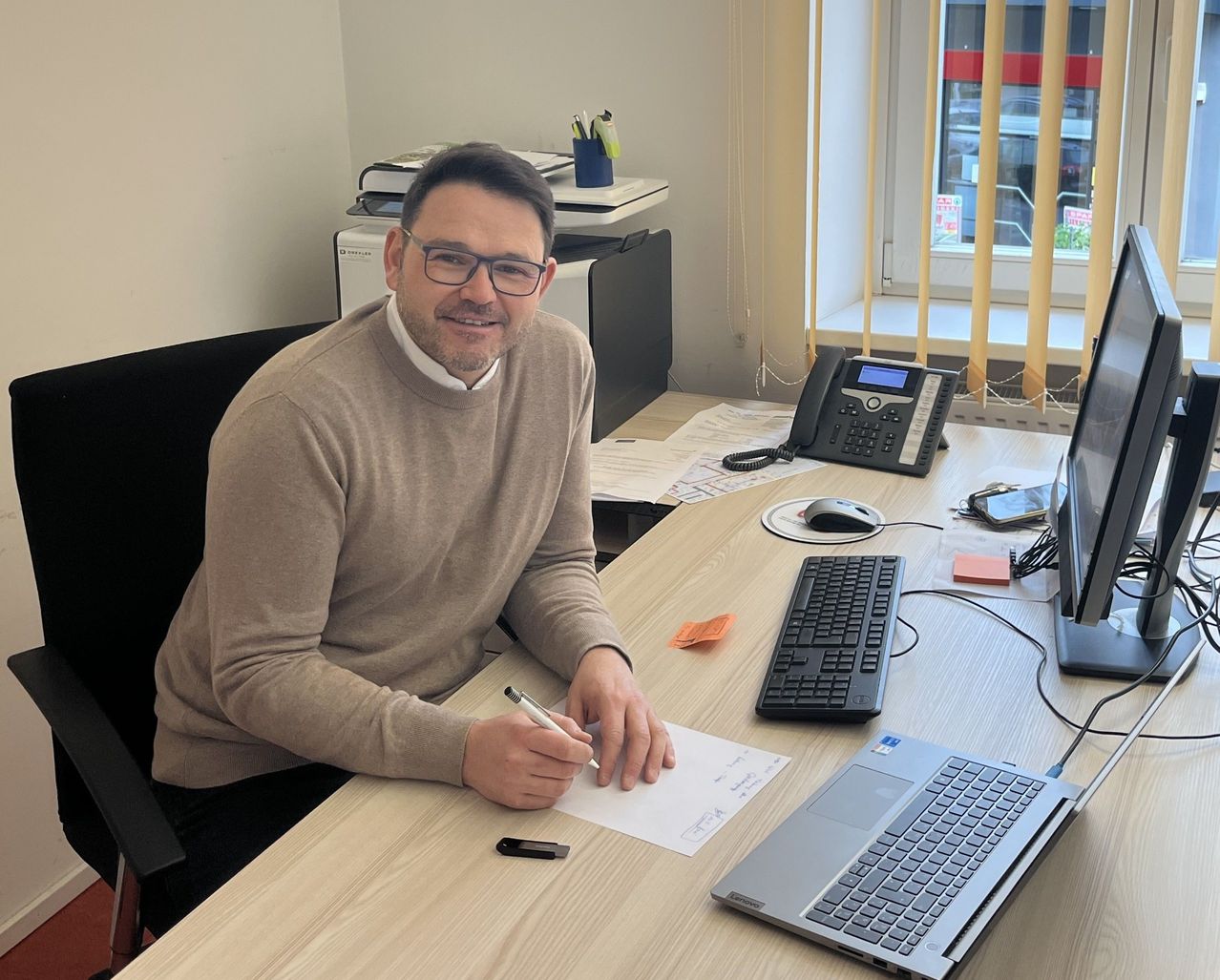 Ein Mann sitzt in einem Büro an einem Schreibtisch und hält einen Stift in der Hand, während er auf einem Blatt Papier schreibt. Auf dem Schreibtisch befinden sich ein Laptop, eine Tastatur und ein Telefon. Hinter ihm ist ein Fenster mit Jalousien.