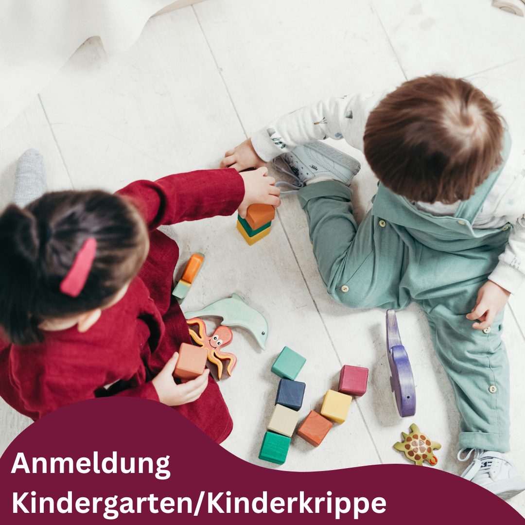 Zwei Kinder spielen auf dem Boden mit Spielzeug. Ein Mädchen in einem roten Shirt spielt mit einem Jungen in Grün. Der Textüberlagerung lautet 'Anmeldung Kindergarten/Kinderkrippe'.