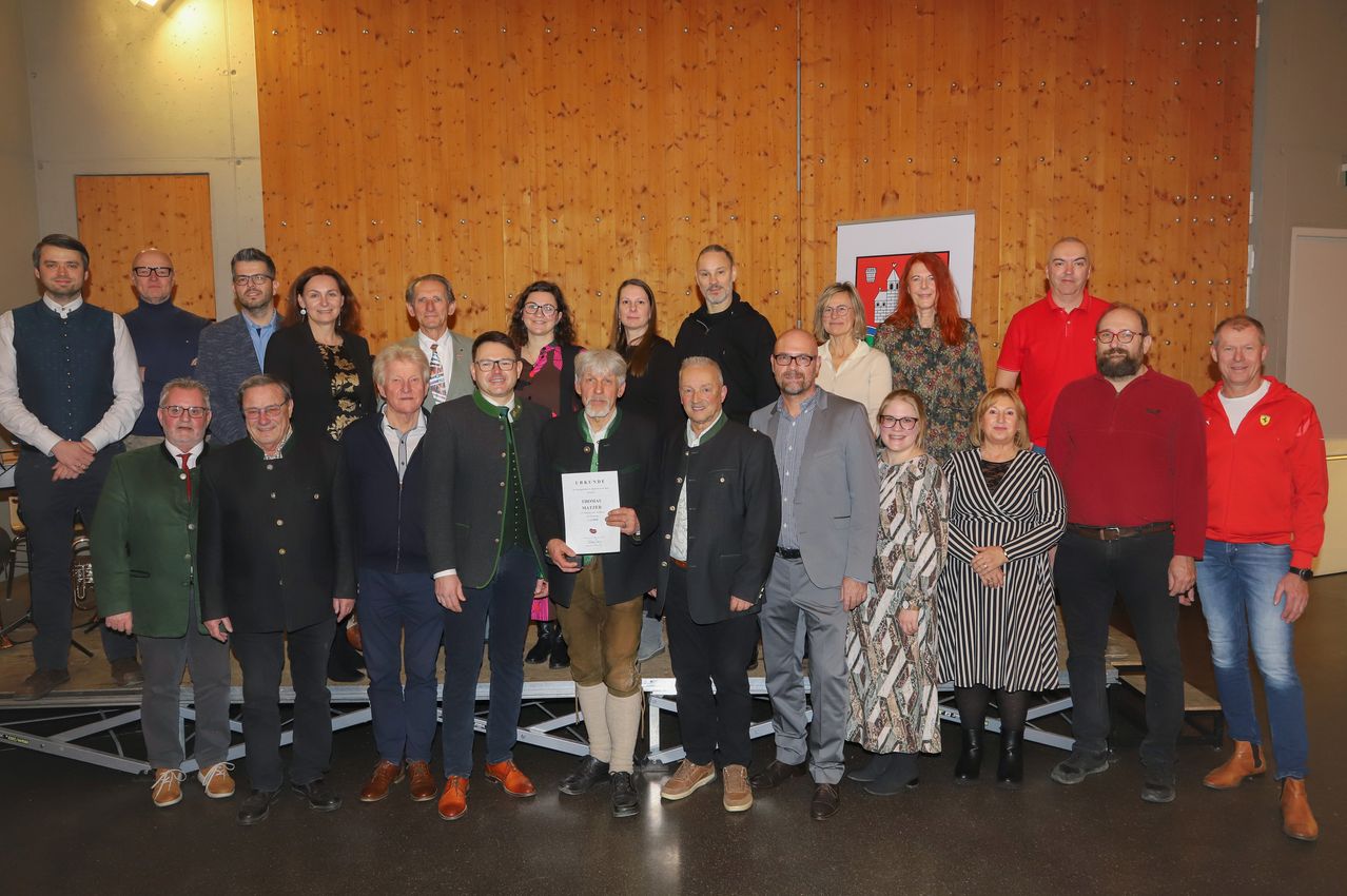 Eine Gruppe von Menschen, möglicherweise ein Team, posiert für ein Foto in einem Raum mit einer Holzwand. Ein Mann in der Mitte hält ein Zertifikat.