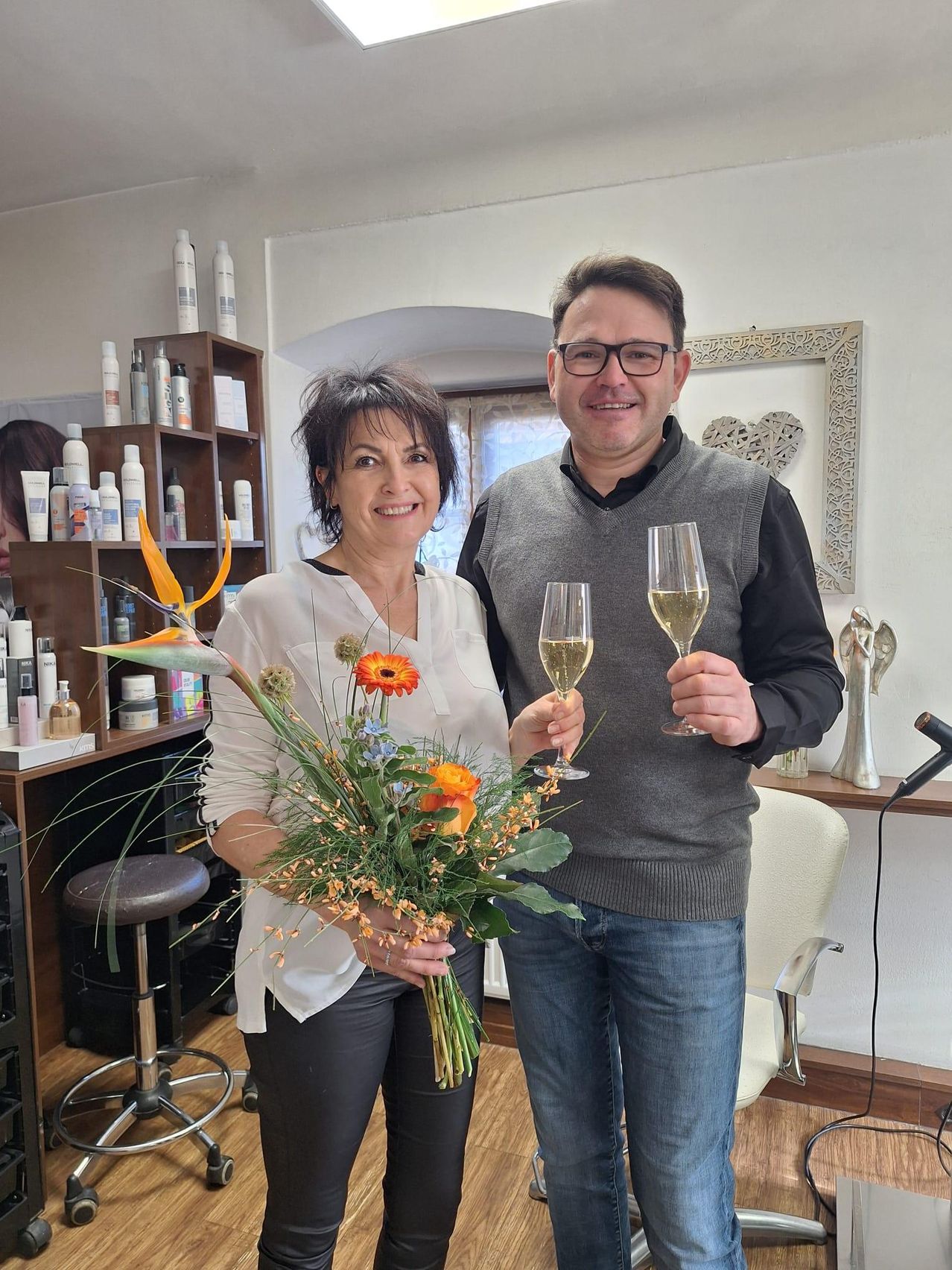 Ein lächelnder Mann und eine Frau halten Weinglas und Blumenstrauß und stehen in einem Salon mit Regalen voller Produkte.