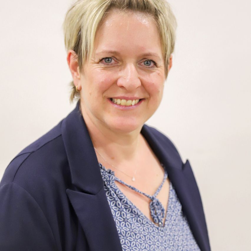 Eine Frau mit kurzen blonden Haaren lächelt und posiert für ein Foto. Sie trägt einen marineblauen Blazer über einem hellblauen Oberteil. Sie hat eine Halskette und Ohrringe. Der Hintergrund ist schlicht.