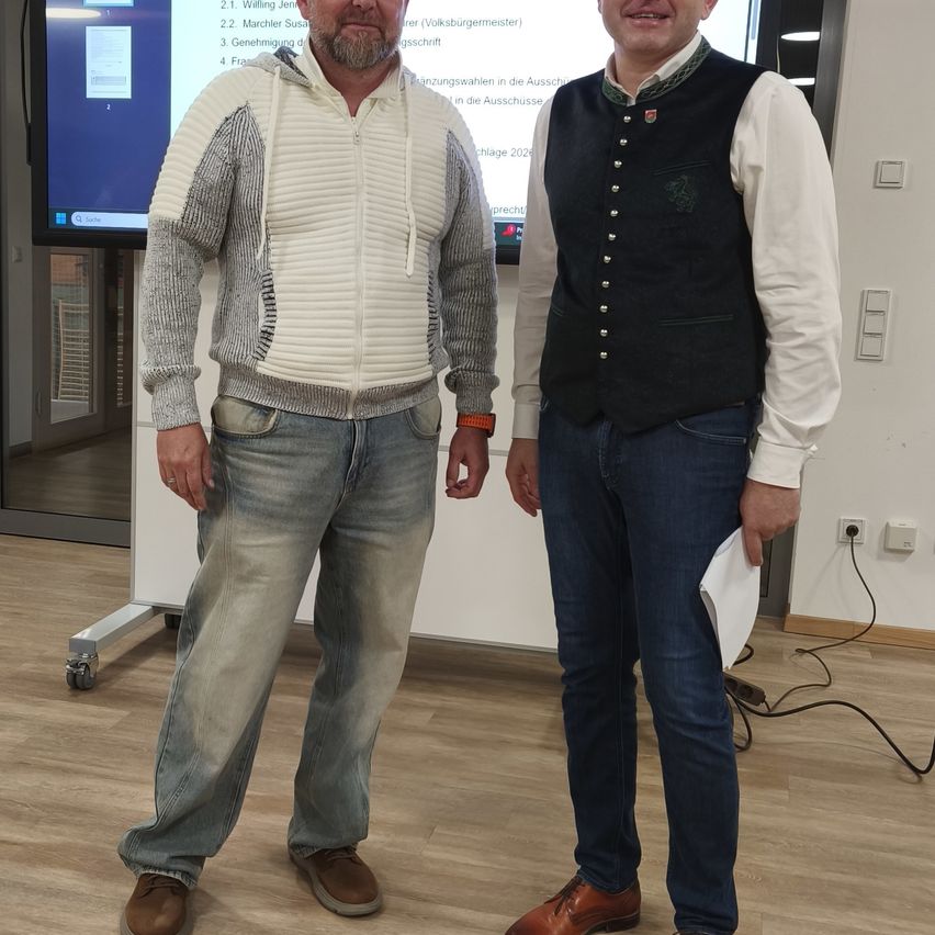 Zwei Männer stehen nebeneinander in einem Raum mit einem Bildschirm im Hintergrund. Ein Mann trägt eine weiße Jacke mit Reißverschluss und der andere trägt eine schwarze Weste über einem weißen Hemd. Sie tragen beide Jeans und Lederschuhe.