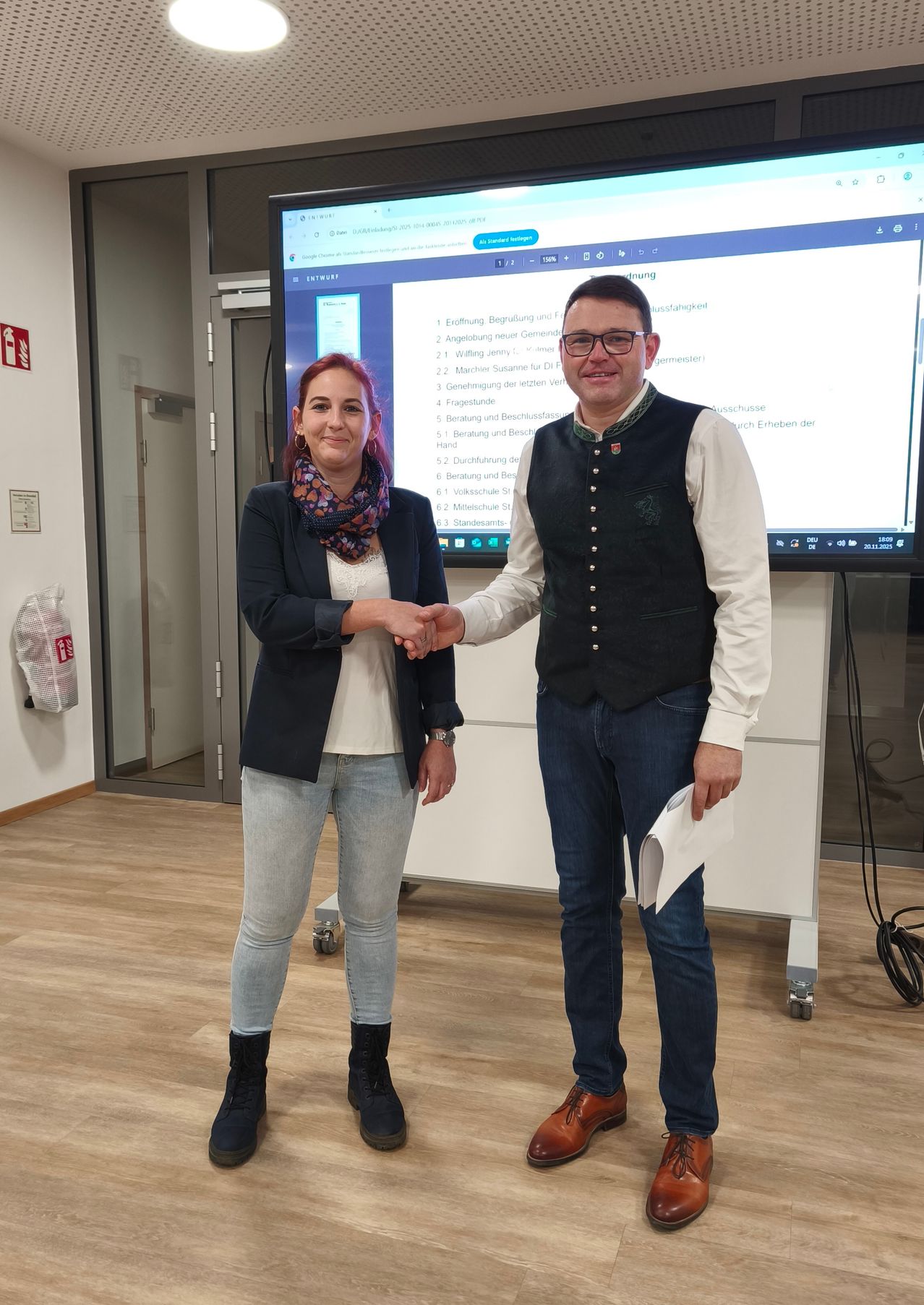 Ein Mann und eine Frau stehen in einem Raum und schütteln Hände. Hinter ihnen ist ein großer Bildschirm, der eine Präsentation zeigt. Der Mann trägt eine Weste und die Frau trägt einen Schal.