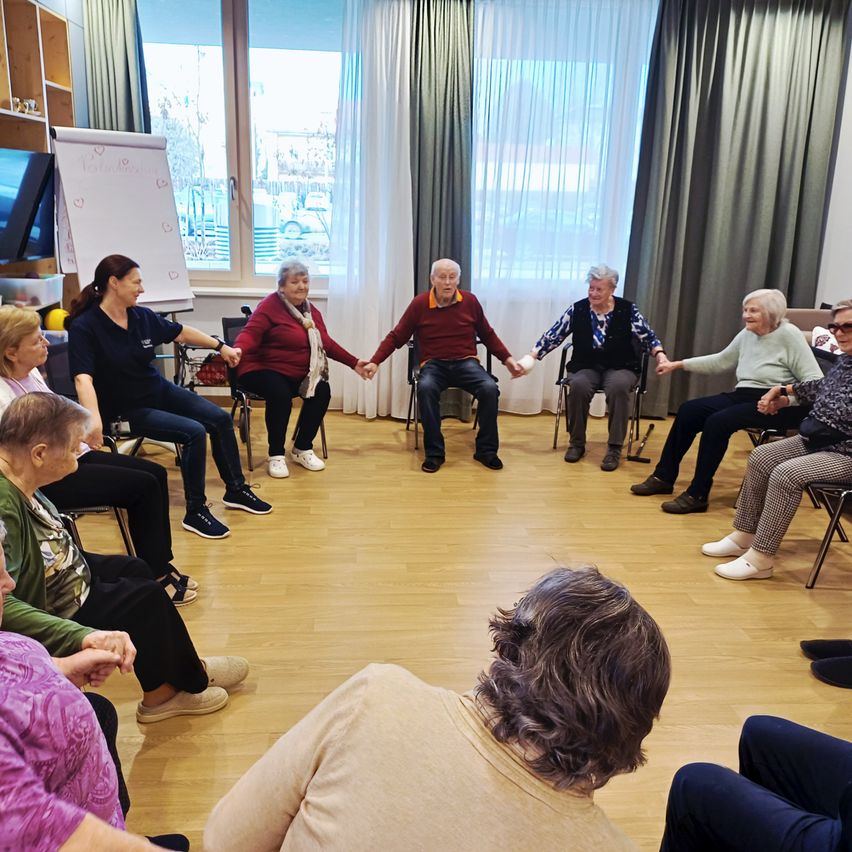 Elderly people are sitting in a circle, holding hands and listening. A woman is leading the activity. The room has wooden floors and curtains. There are two windows with a view of the outside.