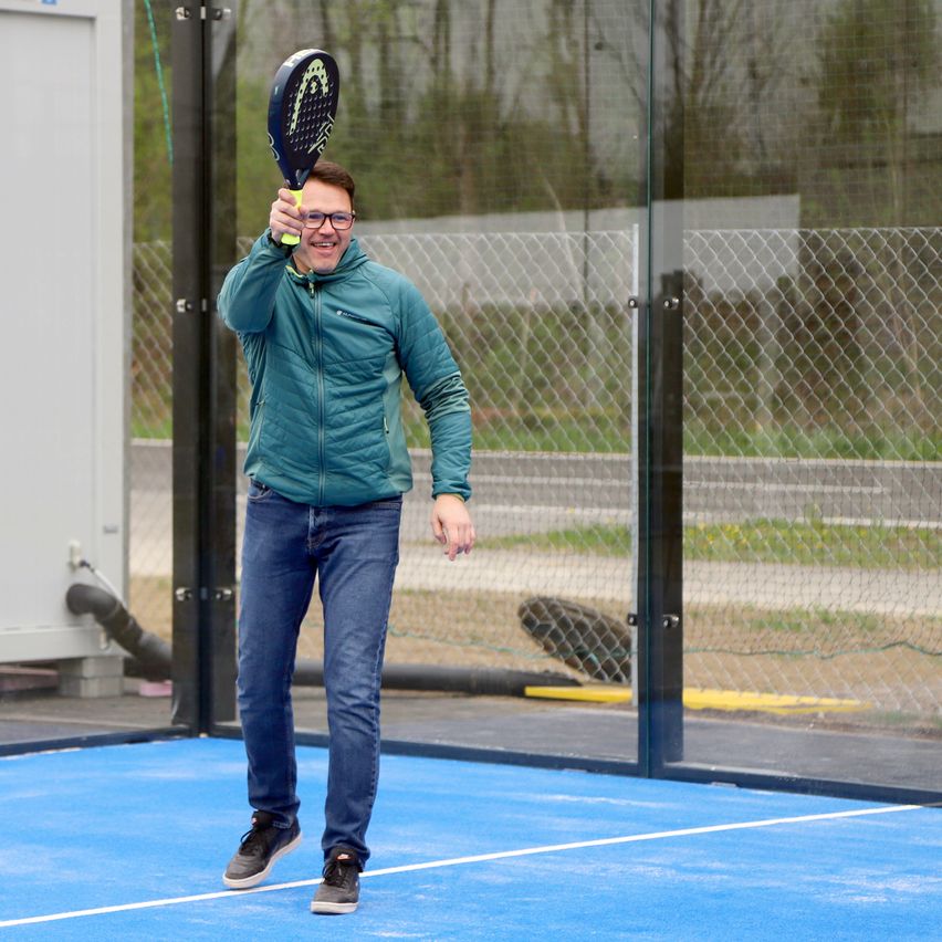 A man holds a paddle tennis racket outdoors on a blue court. He wears a green jacket and blue jeans. Behind him is a glass fence with a chain-link fence.