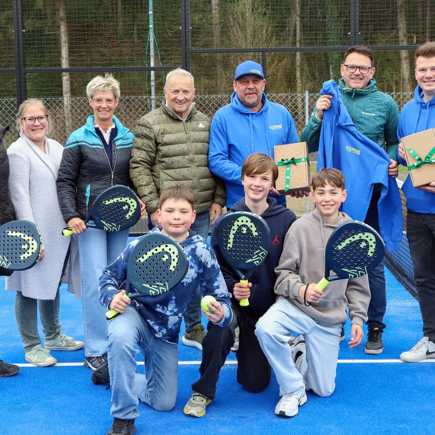 A group of people, including children, stand on a tennis court with blue surfaces, holding rackets and gifts. They are all smiling and posing for a photo.