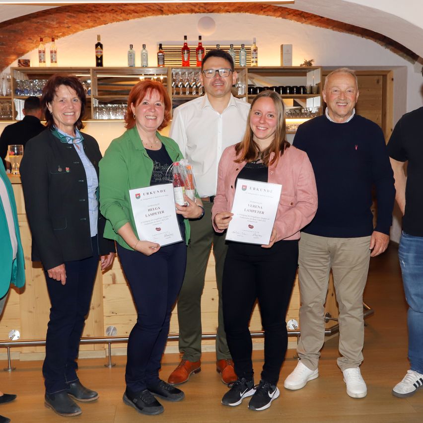 Five adults stand indoors, smiling, holding certificates. One man wears glasses, another a white shirt. The woman in green holds a bag, and another holds a certificate. Behind them, shelves with bottles and glasses are visible.