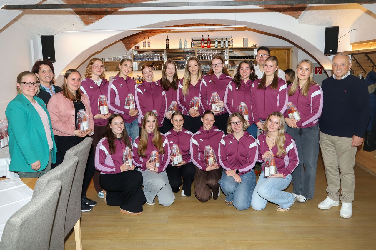 A group of young women in matching uniforms pose for a picture, holding awards and smiling. They are in a room with wooden floors and brick walls.