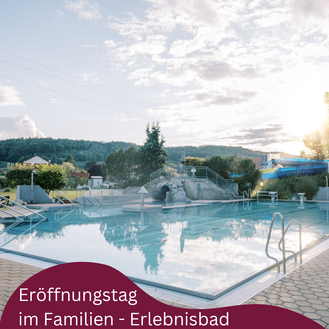 Außenpool mit Rutsche und Eröffnungsschild für Familien, in einem Park mit viel Grün und Bergen im Hintergrund.
