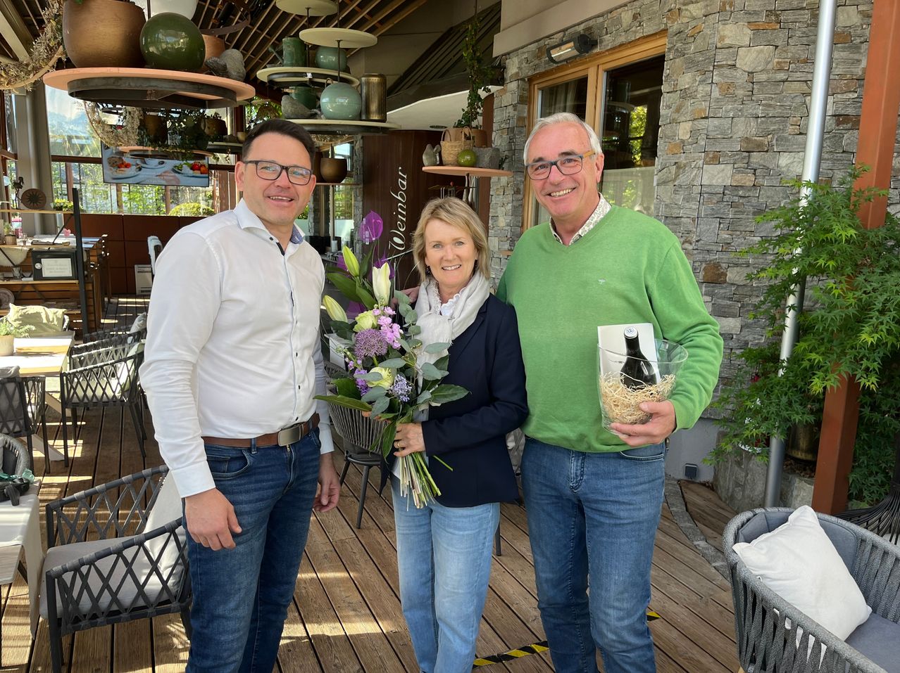 Drei Menschen stehen lächelnd auf einer Holzterrasse. Ein Mann in einem weißen Hemd und Jeans hält Blumen, und ein Mann in einem grünen Pullover hält eine Flasche. Eine Frau in einem blauen Mantel und Schal steht zwischen ihnen.