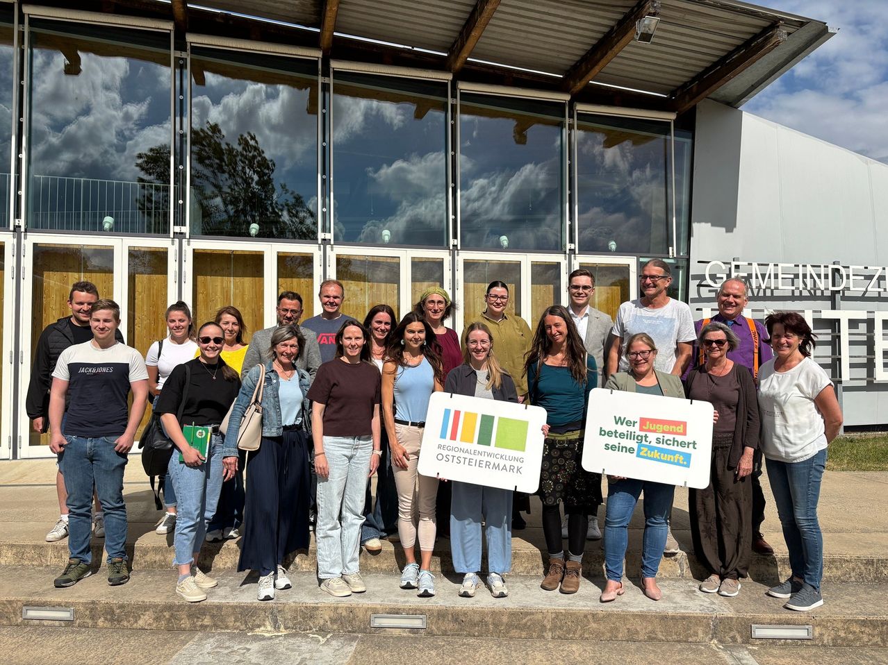 Eine Gruppe von Menschen posiert für ein Foto vor einem Gebäude mit großen Glasfenstern. Einige halten Schilder in der Hand und einige tragen eine Brille. Der Himmel ist bewölkt.
