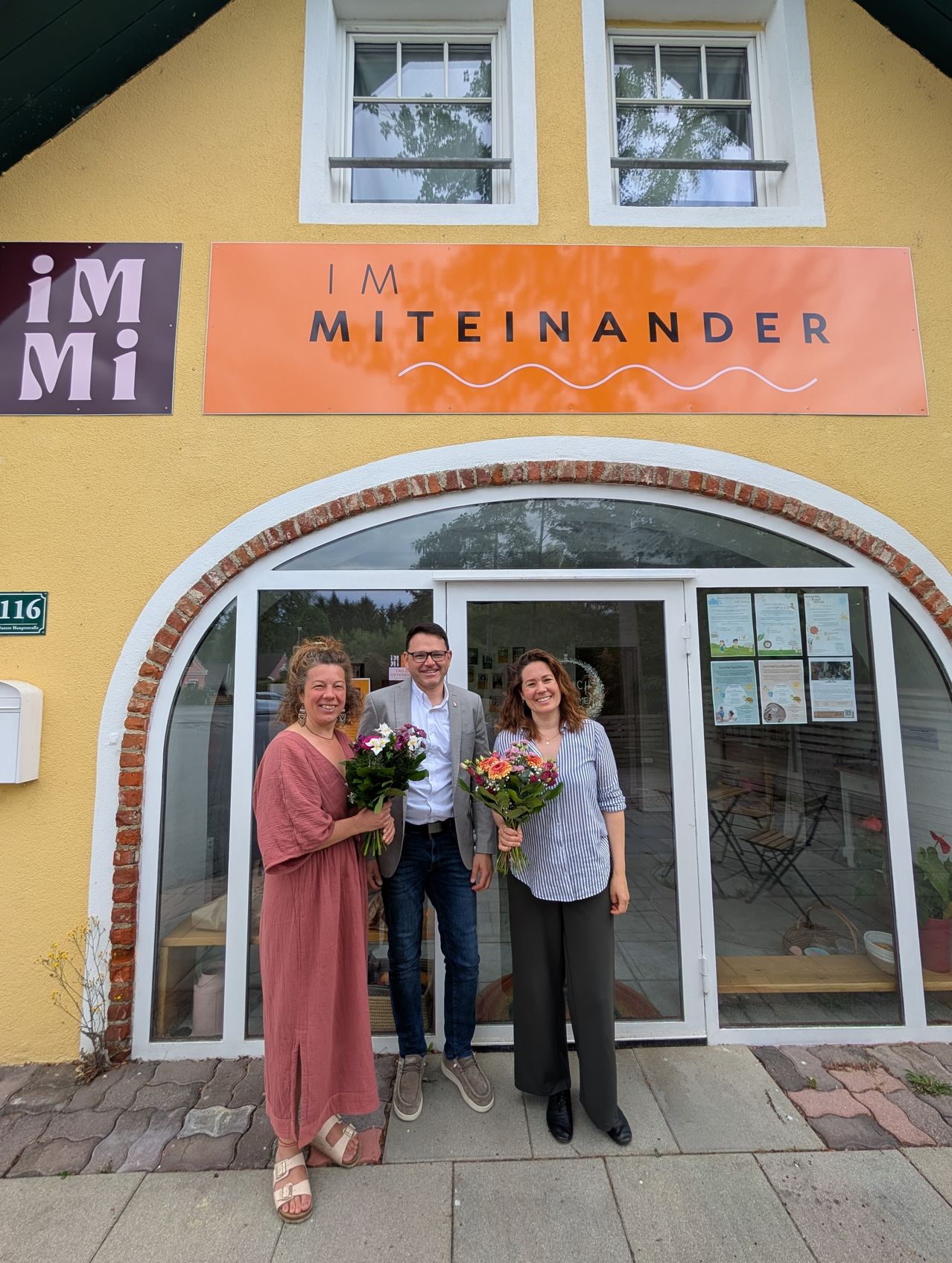 Drei Menschen stehen vor einem Geschäft mit einem orangefarbenen Schild, das IM MITEINANDER steht. Die Frau auf der linken Seite hält Blumen, der Mann in der Mitte lächelt und die Frau auf der rechten Seite lächelt.