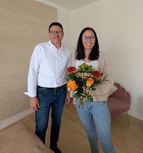 Ein Mann und eine Frau stehen in einem Zimmer, lächeln und halten einen Blumenstrauß. Die Frau trägt eine Brille und einen Pullover. Hinter ihnen ist eine strukturierte Wand und ein rosa Stuhl.