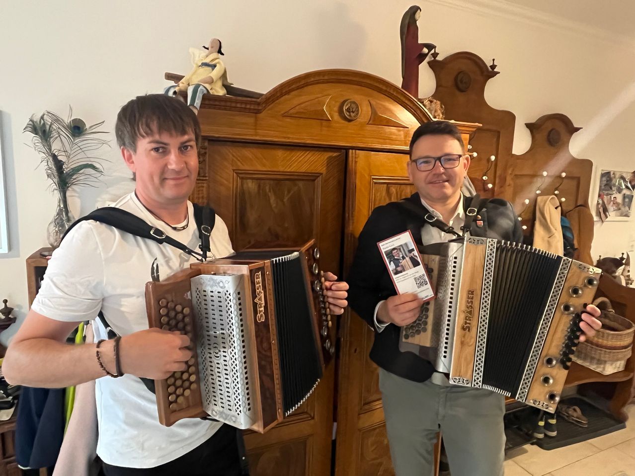 Zwei Männer stehen nebeneinander vor einem hölzernen Schrank und halten beide Akkordeons. Der Mann auf der linken Seite trägt ein weißes T-Shirt und hat einen schwarzen Riemen über der Schulter. Der Mann auf der rechten Seite trägt einen schwarzen Anzug und hält eine Karte.