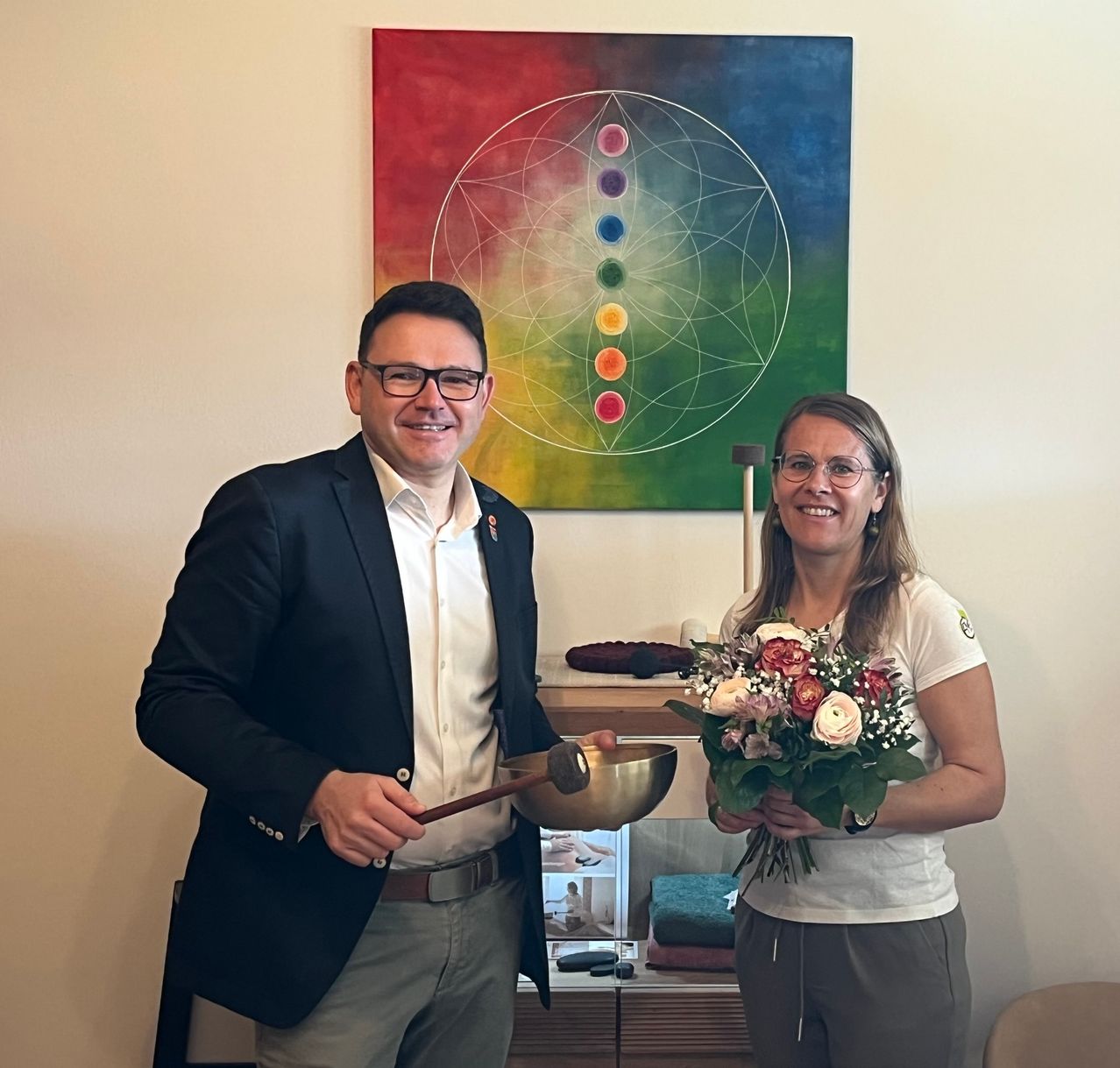 Ein Mann und eine Frau stehen lächelnd zusammen in einem Raum, mit einem Blumenstrauß und einer Klangschale. Ein farbenfrohes Gemälde mit einem Mandala-Muster befindet sich an der Wand hinter ihnen.