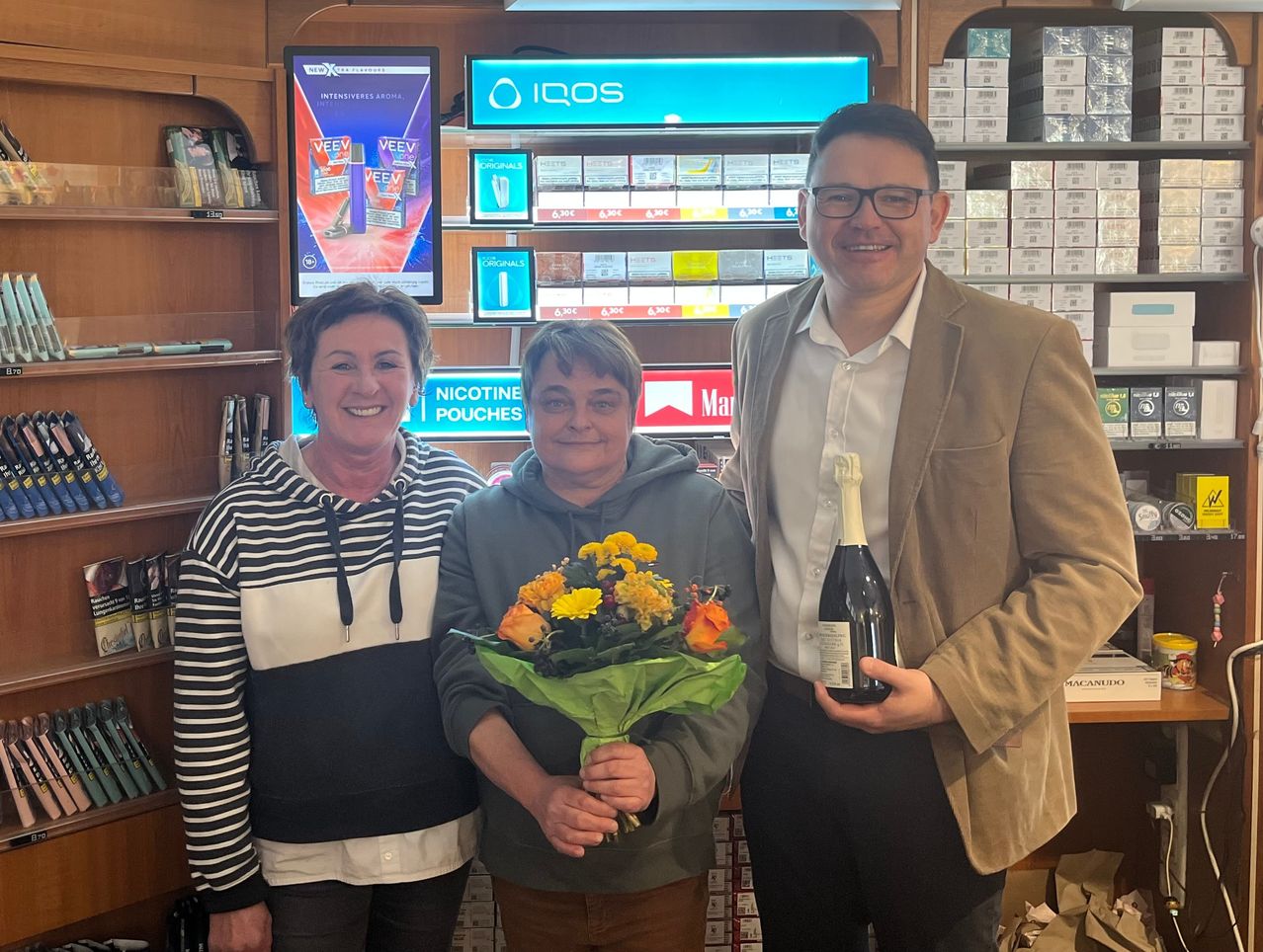 Drei Personen stehen in einem Geschäft mit Produkten im Hintergrund. Der Mann hält eine Flasche und lächelt, während die Frau mit einem Blumenstrauß lächelt. Dahinter sind Regale mit verschiedenen Produkten zu sehen.