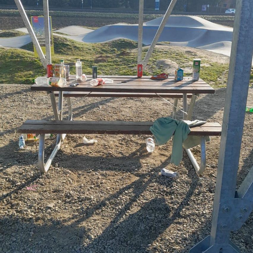 Ein Picknicktisch in einem Park ist mit leeren Dosen, Flaschen und einem grünen Tuch übersät. In der Nähe befindet sich ein Skatepark mit Rampen.