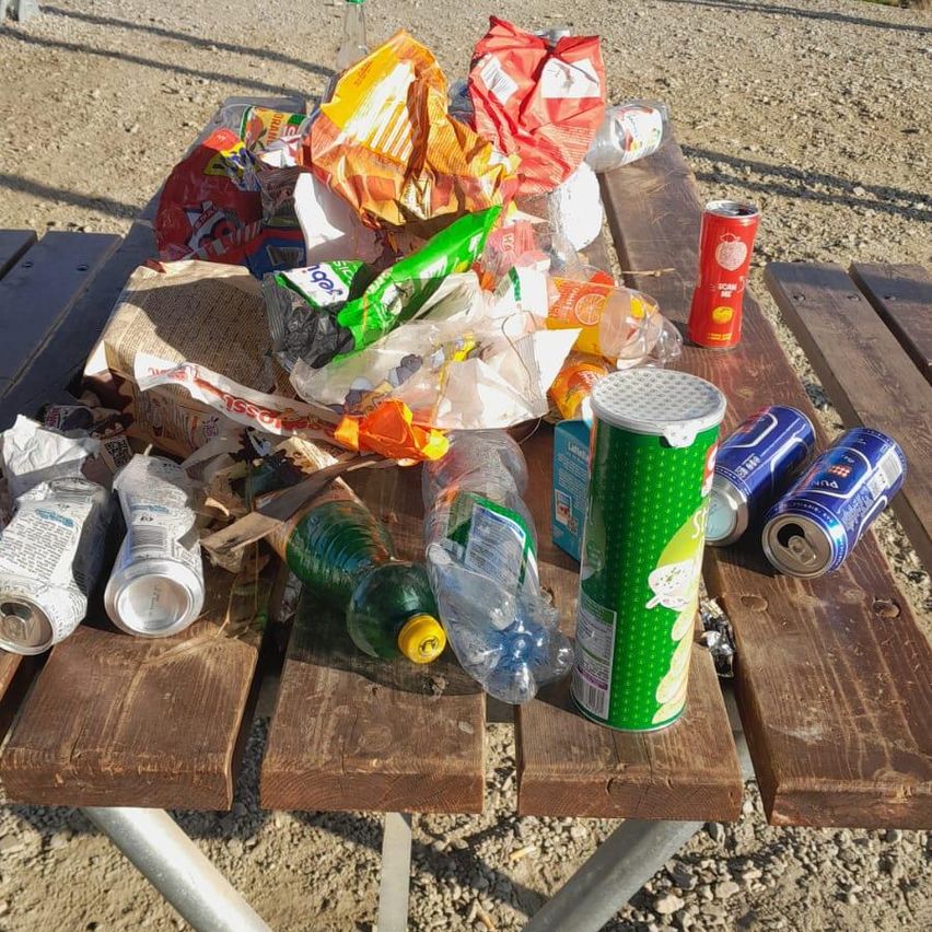 Ein Holz-Picknicktisch, gefüllt mit verschiedenem Müll, darunter leere Plastikflaschen, Dosen, Verpackungen und anderer Abfall.