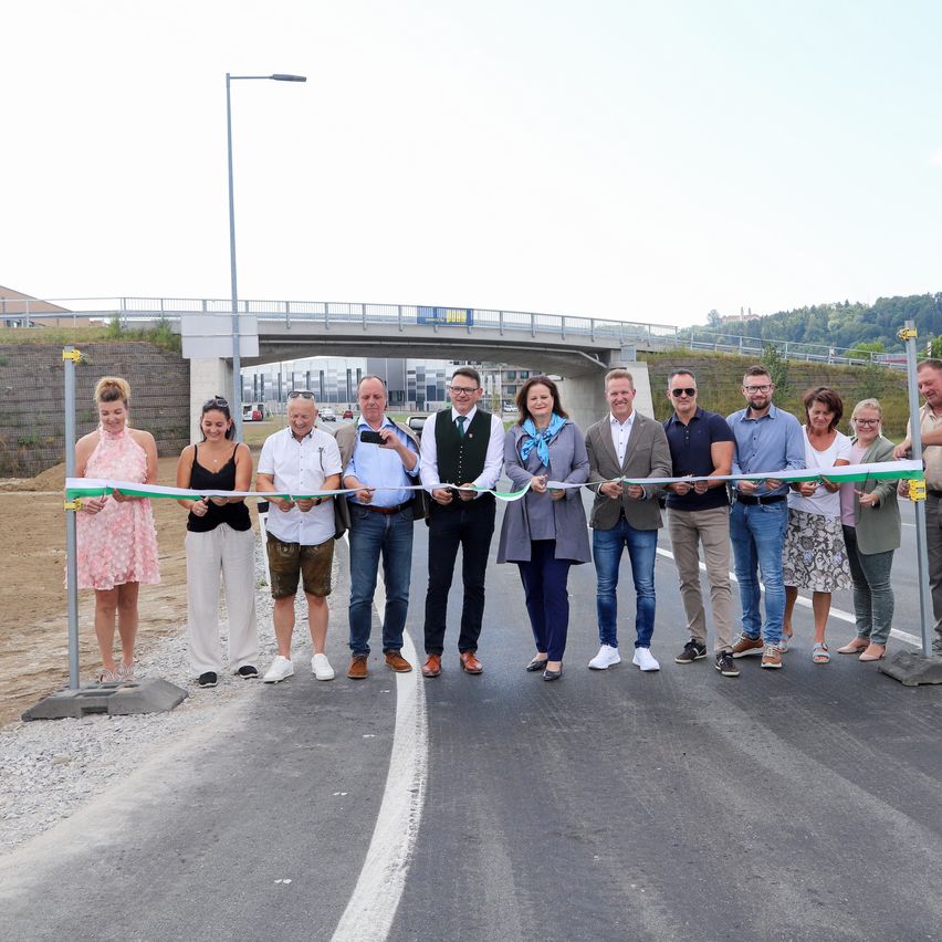 Eine Gruppe von Menschen steht auf einer neuen Straße und hält ein Band, um die Eröffnung zu feiern. Sie lächeln und posieren für ein Foto. Dahinter befindet sich eine Brücke und ein Gebäude.