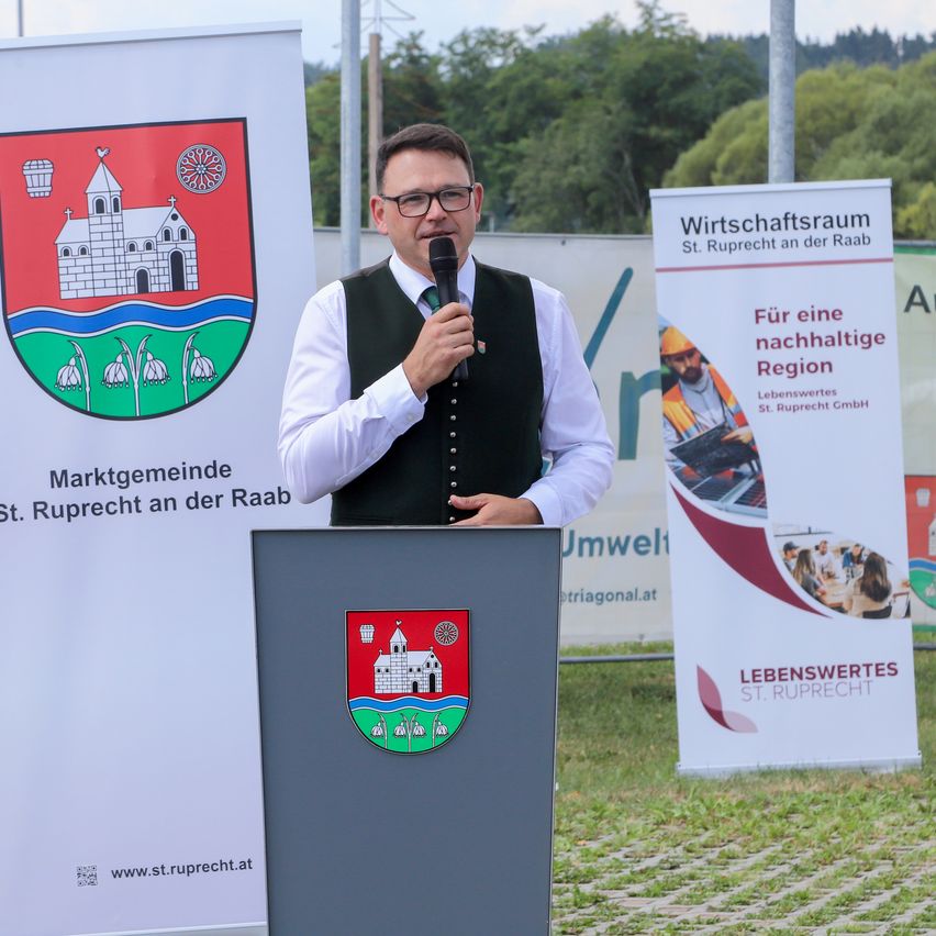 Ein Mann spricht in ein Mikrofon an einem Rednerpult mit einem Schild hinter ihm, das 'Marktgemeinde St. Ruprecht an der Raab' steht.