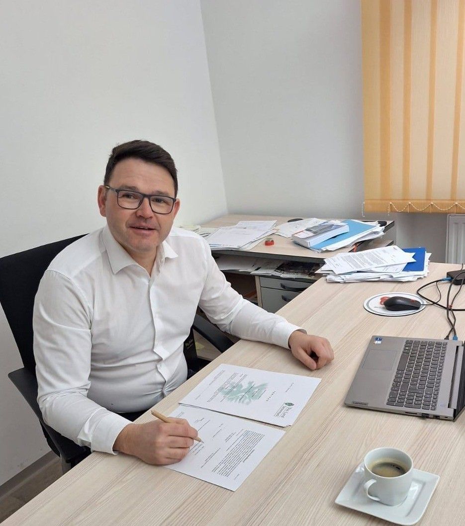 Ein Mann in einem weißen Hemd sitzt an einem Schreibtisch mit Papieren, einem Laptop und einer Tasse Kaffee. Er hält einen Stift und schaut in die Kamera.