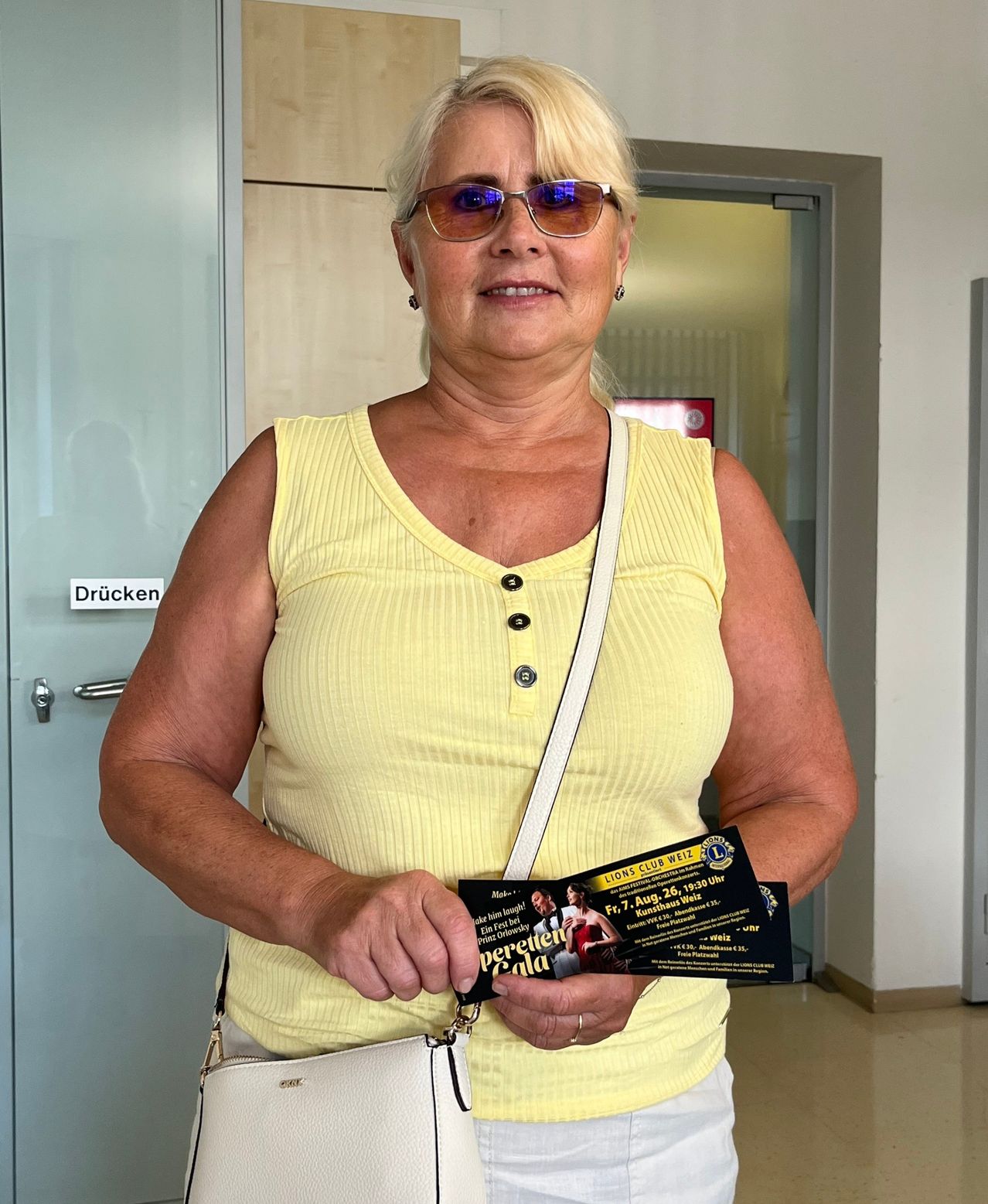 Eine ältere Frau mit blonden Haaren trägt ein gelbes ärmelloses Oberteil und Sonnenbrille und hält ein Ticket für eine Lions Club Veranstaltung.