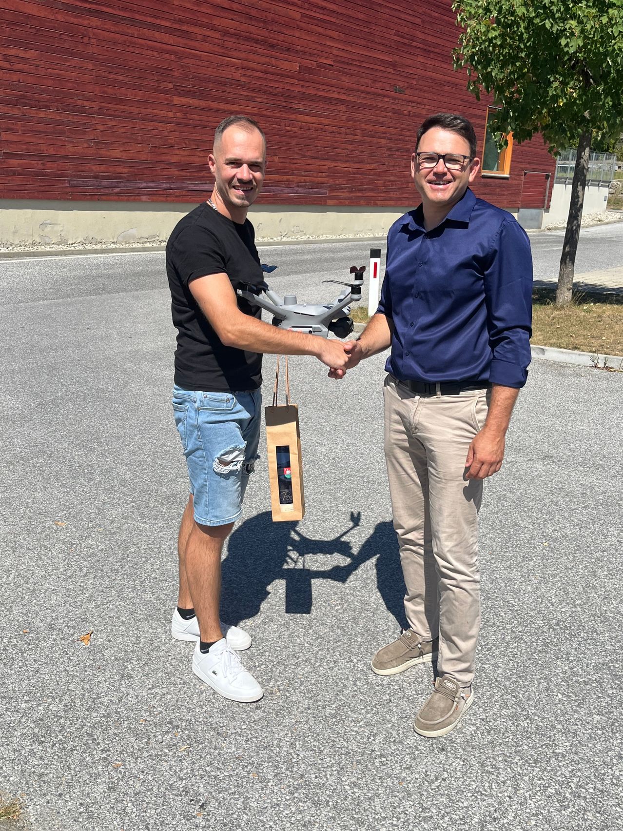 Two men are shaking hands in front of a building. One is holding a drone and a bag.