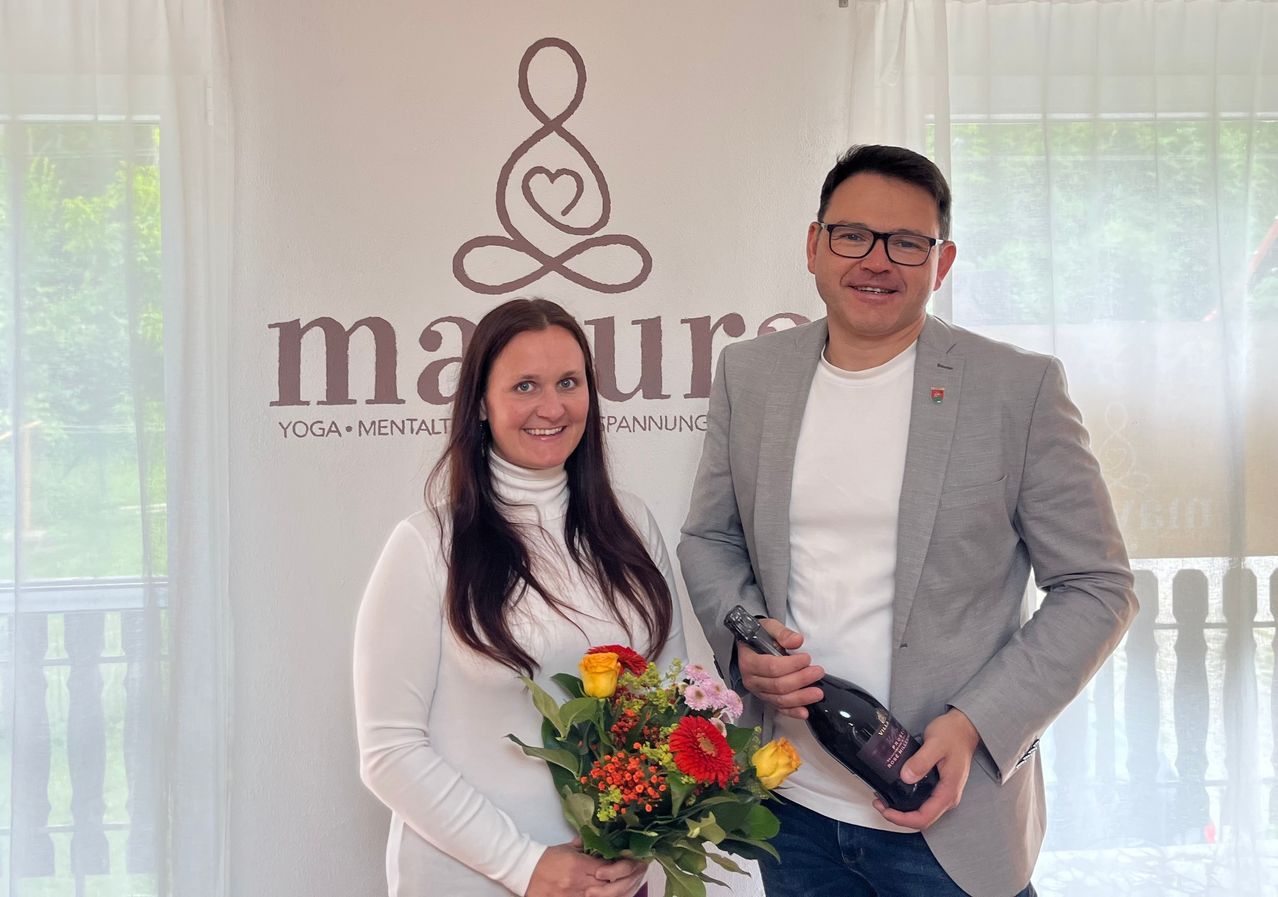 Ein Mann und eine Frau stehen nebeneinander und halten einen Blumenstrauß und eine Flasche Wein. Sie lächeln und scheinen ein Foto vor einer Wand mit Logo zu machen.
