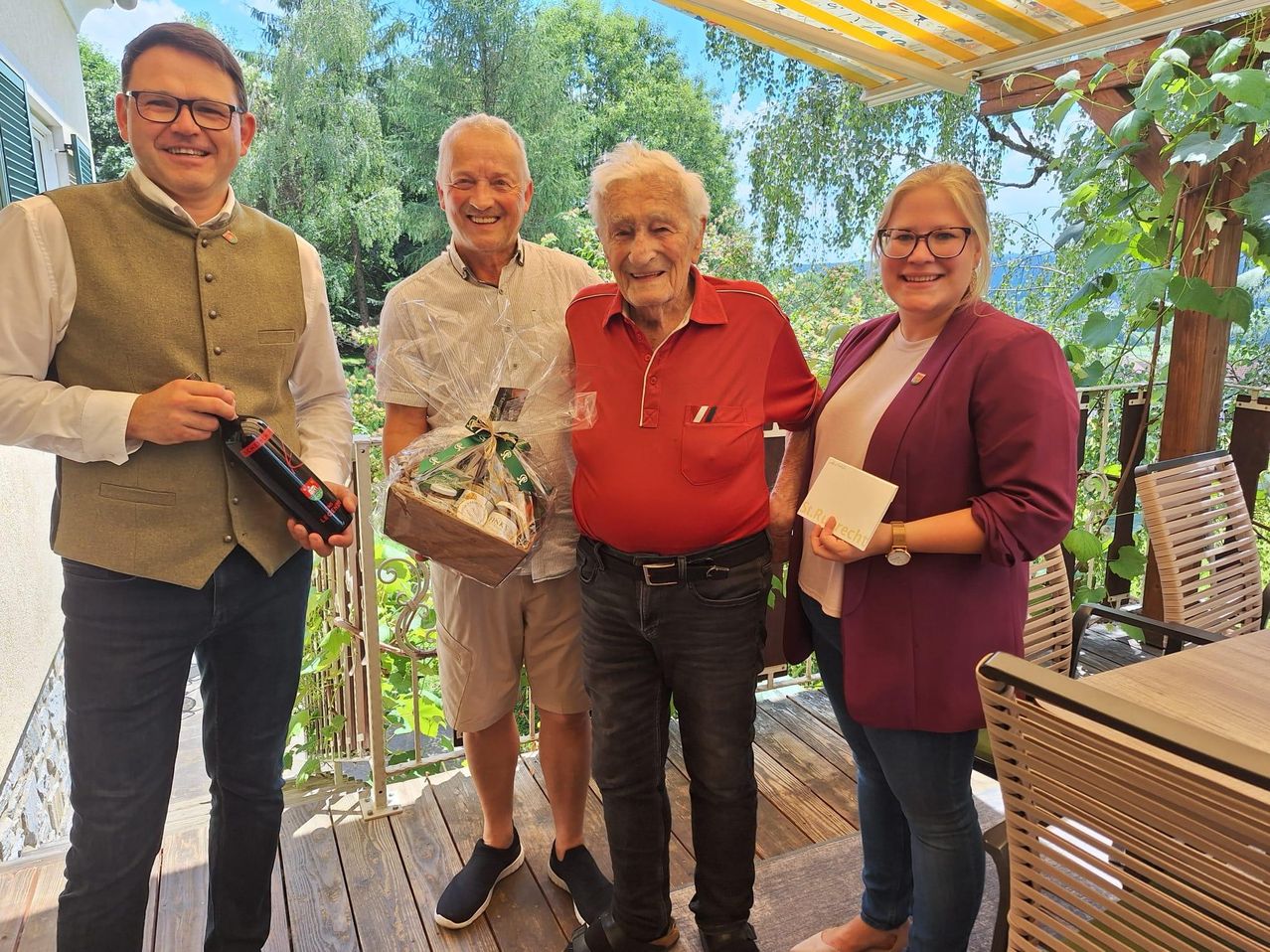 Vier Personen auf einer Terrasse, ein älterer Mann hält ein Geschenk, ein Mann hält eine Flasche und zwei andere lächeln. Bäume und ein Zaun im Hintergrund.