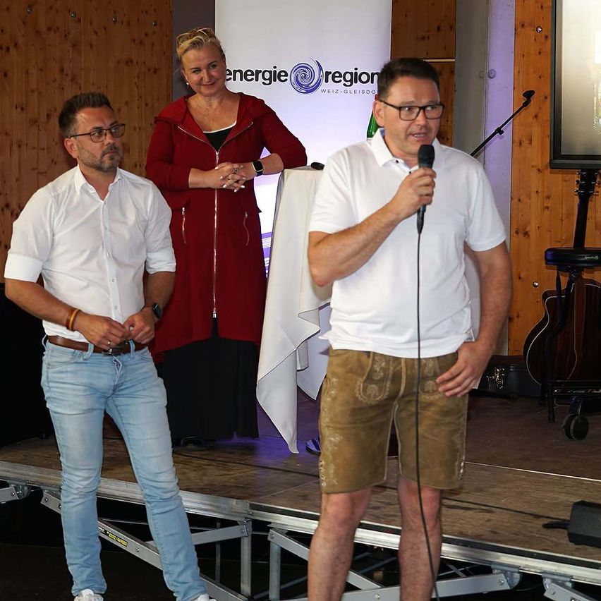 Drei Personen stehen auf einer Bühne, zwei Männer und eine Frau. Der Mann rechts spricht in ein Mikrofon, während der andere Mann und die Frau hinter ihm stehen.