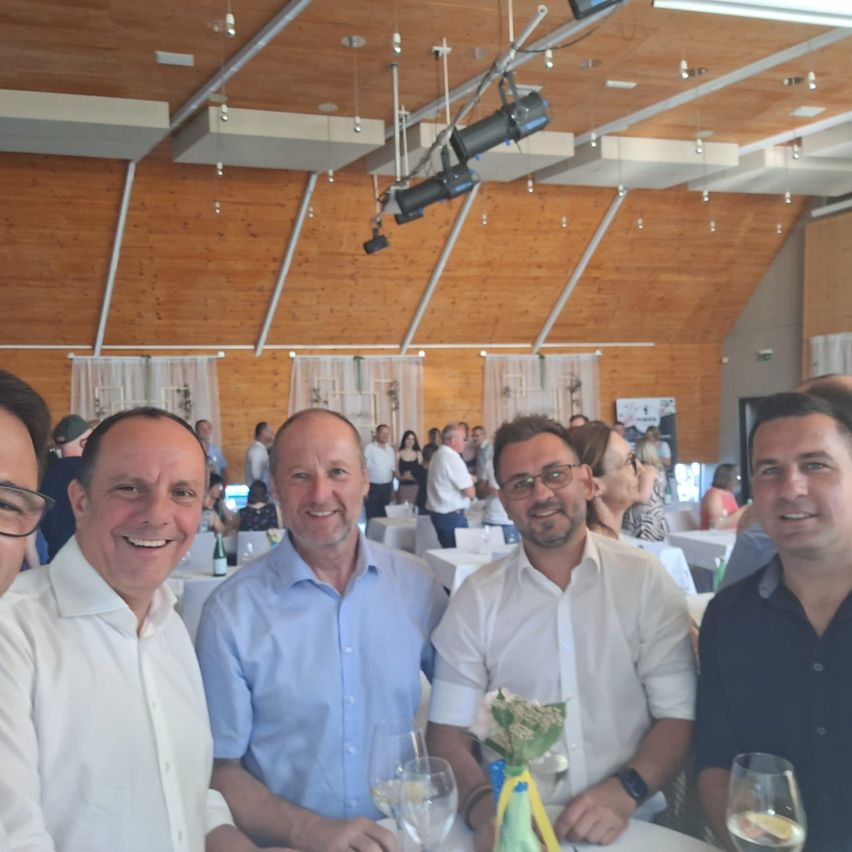 Vier Männer lächeln und posieren für ein Foto bei einer Indoor-Veranstaltung. Sie sitzen an einem Tisch mit Weingläsern und einem Blumen-Zentrem. Der Raum hat Holzwände und Decken mit Scheinwerfern.