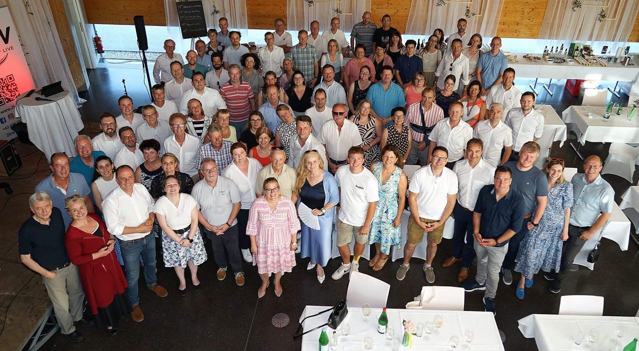 Eine große Gruppe von Menschen, wahrscheinlich ein Team, posiert für ein Foto bei einer Veranstaltung. Die Umgebung scheint drinnen zu sein, mit Tischen, Stühlen und einer Tafel sichtbar.