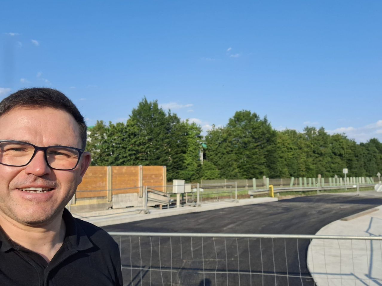 A man stands near a newly paved road under a clear blue sky, with a wooden barrier and greenery in the background.