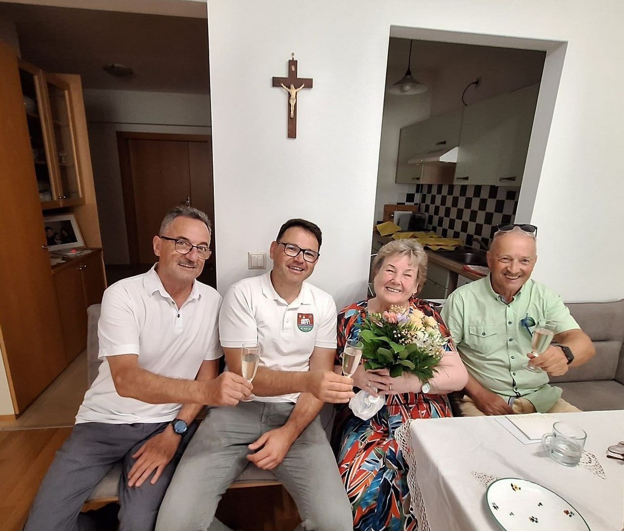 Four people sit in a room. Three men wear white shirts. A woman holds a bouquet. They all smile and hold glasses of wine. A cross is on the wall.