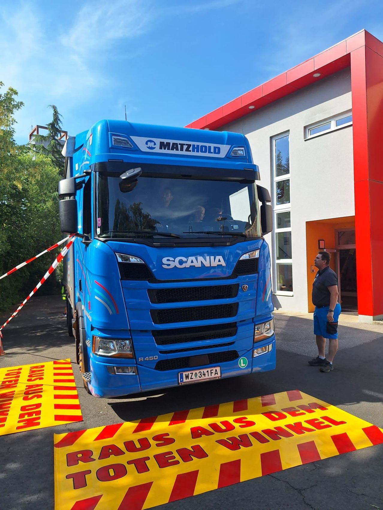 A blue Scania truck with the brand name Matzhold is parked in front of a building. A man stands nearby, wearing blue shorts.
