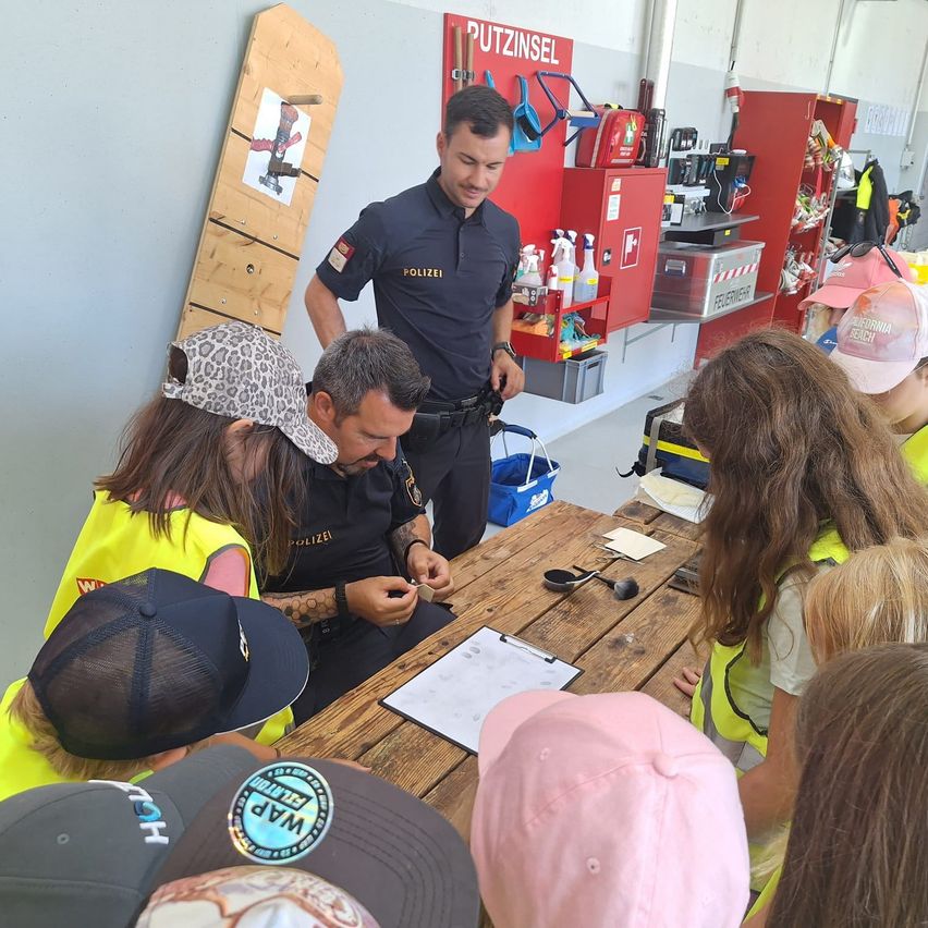 A group of children in yellow vests are gathered around a table with a police officer and another man in a workshop.