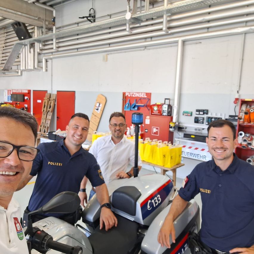 Four men smile in a garage, three in blue police uniforms and one in white. A motorcycle is in the foreground with the number 133 on it. Behind them, there are red doors and a table with yellow bags.