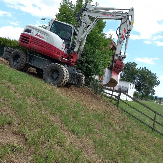 Ein roter und weißer Bagger gräbt auf einem Grashang Erde aus, mit einem Haus und Bäumen im Hintergrund.