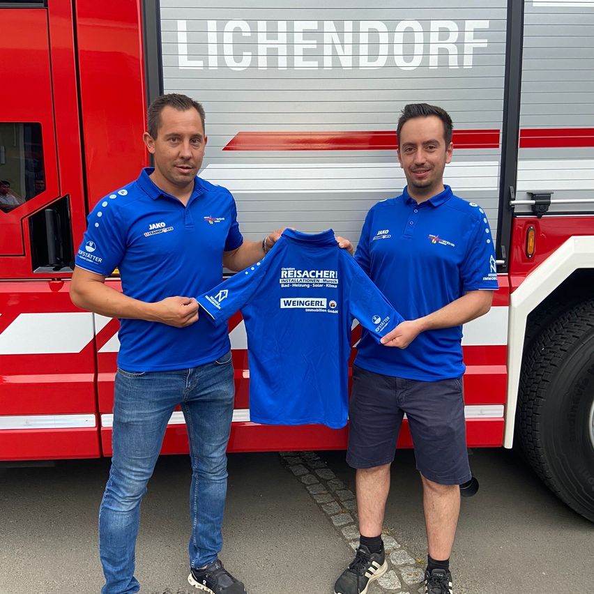 Zwei Männer stehen vor einem roten Feuerwehrauto, einer hält ein blaues Shirt mit dem Namen 'REISACHER'. Beide Männer tragen blaue Poloshirts und Jeans.