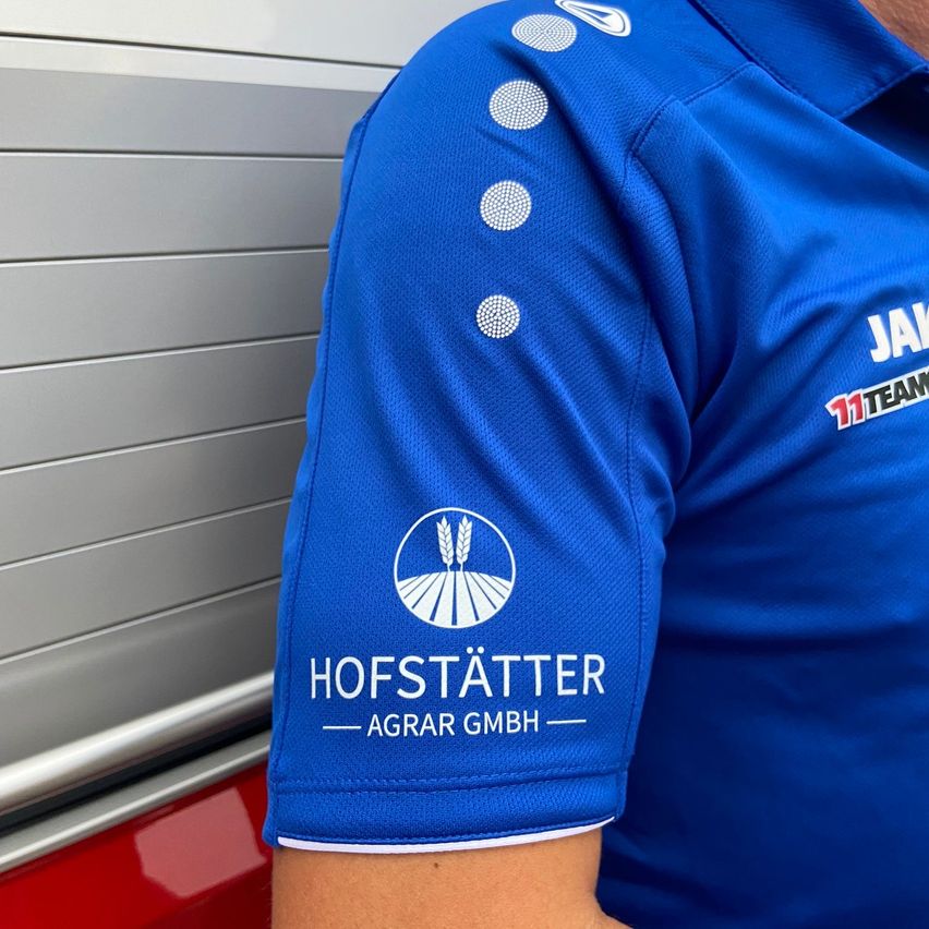 Ein blaues Poloshirt mit dem Logo 'HOFSTATTER AGRAR GMBH' und einem Weizenlogo auf dem Schulterbereich ist abgebildet. Das Wort 'JAK' befindet sich oben rechts.
