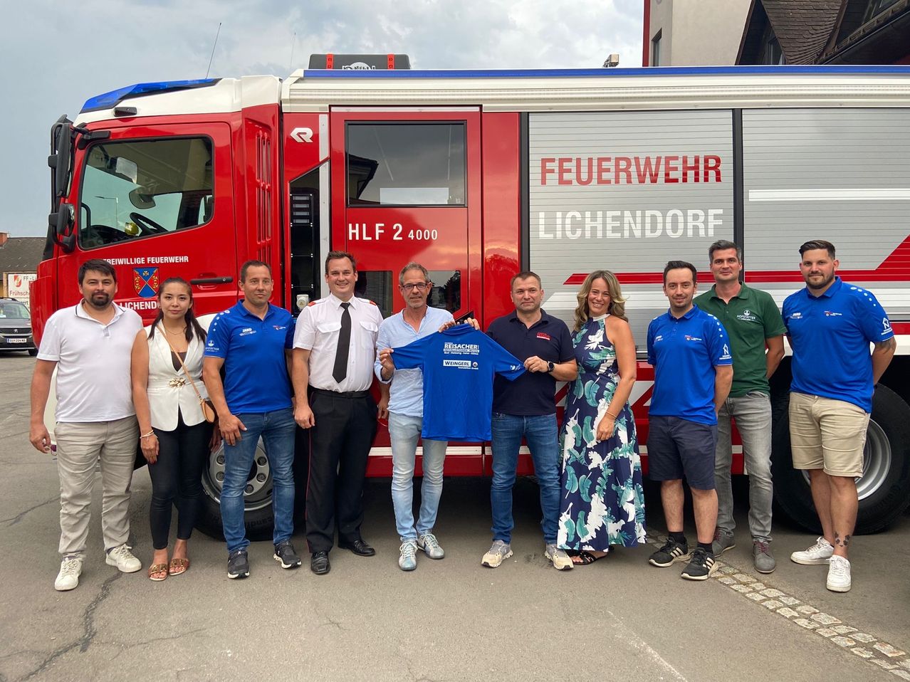 Eine Gruppe von Menschen posiert vor einem roten Feuerwehrauto. Das Auto trägt die Aufschrift 'Feuerwehr' und 'H.L.F 2 4000'. Zwei Männer halten ein blaues Trikot.