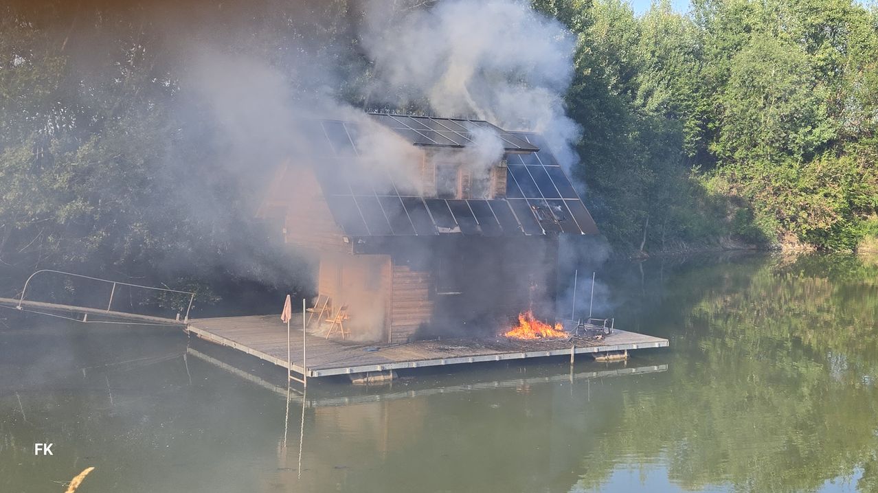 Ein Holzhaus auf einem Seesteg steht in Flammen. Rauch dringt aus den Fenstern, und das Feuer ist vorne sichtbar.