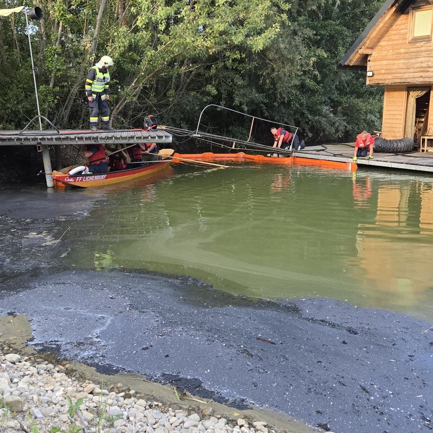 Feuerwehrleute in gelben Westen stehen auf einer Anlegestelle am Fluss. Ein kleines Boot ist in der Nähe angedockt. Im Hintergrund befinden sich Bäume und ein Holzhaus.